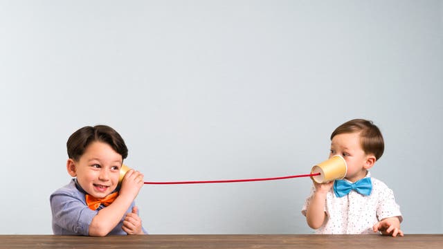 Zwei Kinder sitzen an einem Tisch und spielen mit einem selbst gemachten Telefon aus Pappbechern und einer Schnur. Das Kind links hält einen Becher ans Ohr und lächelt, während das Kind rechts in den anderen Becher spricht. Beide tragen bunte Fliegen. Der Hintergrund ist neutral.