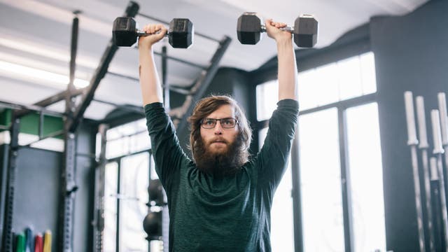 Ein Mann mit Brille und Bart hebt zwei Kurzhanteln über den Kopf in einem Fitnessstudio. Im Hintergrund sind Fitnessgeräte und große Fenster zu sehen. Die Szene vermittelt eine konzentrierte Trainingsatmosphäre.