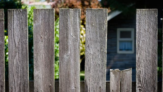 Holzzaun mit einer abgebrochenen Latte, der den Blick auf ein Haus mit Garten dahinter frei gibt.