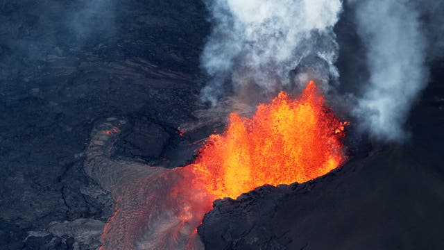 Ein aktiver Vulkan, der glühende Lava und Rauch in die Luft schleudert. Die Lava fließt über die dunkle, erkaltete Oberfläche des Vulkans. Keine Menschen sind im Bild zu sehen.