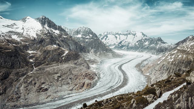 Eine weite Landschaftsaufnahme eines Gletschers, der sich durch ein Tal zwischen schneebedeckten Bergen schlängelt. Der Himmel ist leicht bewölkt, und die Berge zeigen felsige und schneebedeckte Flächen. Der Gletscher erstreckt sich in die Ferne und bildet eine markante, helle Linie in der Mitte des Bildes. Die Szene vermittelt eine ruhige, majestätische Atmosphäre.