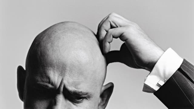 Ein Schwarz-Weiß-Foto zeigt eine Person, die sich nachdenklich den Kopf kratzt. Der Fokus liegt auf der Handbewegung und dem Ausdruck der Konzentration. Die Person trägt einen Anzug mit einem gestreiften Muster. Der Hintergrund ist neutral und lenkt nicht vom Hauptmotiv ab.