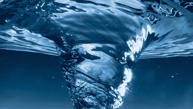 Ein Wirbel aus klarem, blauem Wasser, der sich spiralförmig nach unten bewegt. Die Lichtreflexionen auf der Wasseroberfläche erzeugen glänzende, wellenförmige Muster. 