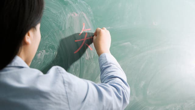Eine Person schreibt mit roter Kreide ein chinesisches Schriftzeichen auf eine grüne Tafel. Die Person ist von hinten zu sehen und trägt ein helles Hemd. Die Tafel zeigt Spuren von vorherigem Schreiben.