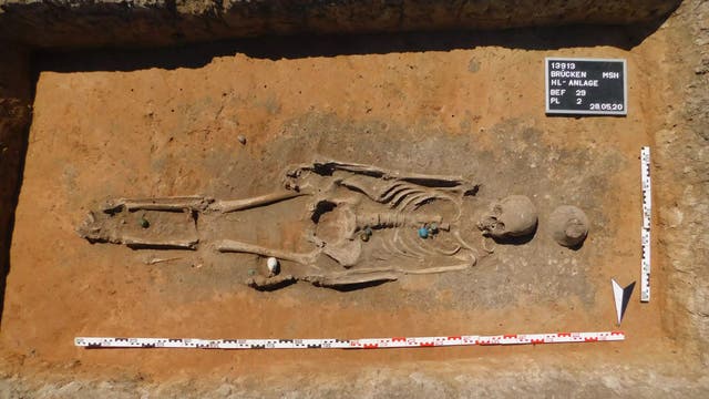 Ein archäologisches Foto zeigt ein freigelegtes menschliches Skelett in einer Grabungsstätte. Das Skelett liegt auf sandigem Boden, umgeben von Messinstrumenten und einer Tafel mit der Aufschrift „13913 Brücken MSH HLC-Anlage BEF 29 PL 2 28.05.20“. Maßstäbe sind am unteren und am rechten Rand des Bildes sichtbar.