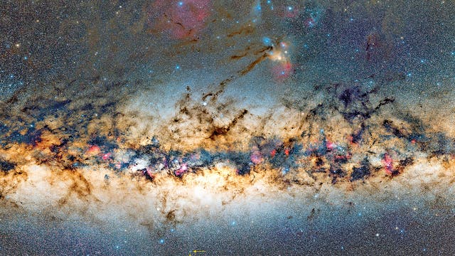 Ein detailliertes Bild der Milchstraße, das den dichten, sternenreichen Streifen der Galaxie zeigt. Dunkle Staubwolken durchziehen das Bild, während bunte Nebel und helle Sterne hervortreten. Ein roter Kreis markiert einen bestimmten Bereich im unteren Teil des Bildes. Die Farben reichen von leuchtendem Blau und Rosa bis zu warmen Gelb- und Brauntönen.