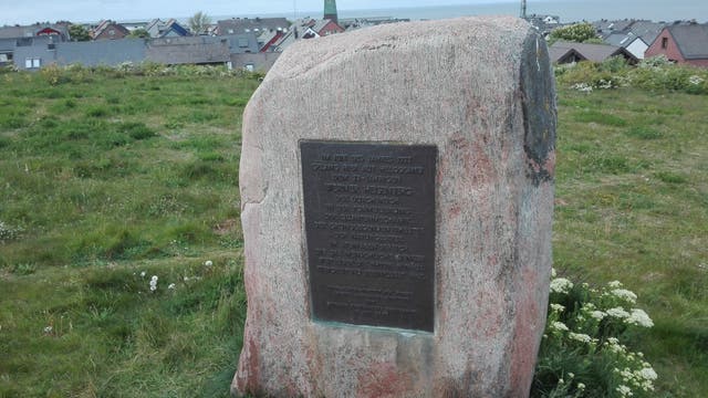 Ein Gedenkstein mit einer Inschrift steht auf einer grünen Wiese. Im Hintergrund sind Häuser mit grauen Dächern und eine Kirche mit einem spitzen Turm zu sehen. Der Himmel ist bewölkt, und im Hintergrund erstreckt sich das Meer.