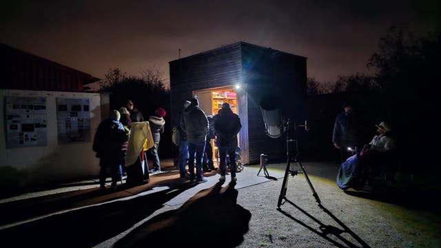 Mehrere Personen halten sich in der Dunkelheit auf einem kleinen Platz vor einer Gerätehütte auf, aus der Licht scheint. Verschiedene Teleskope sind aufgebaut, die von den Anwesenden begutachtet werden.