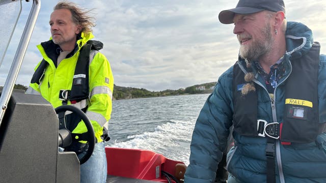 Zwei Personen stehen auf einem Boot vor einem bewölkten Himmel. Beide tragen Rettungswesten; die Person links trägt eine leuchtend gelbe Jacke, die Person rechts eine blaue Jacke und eine Mütze. Vor ihnen ist ein Teil des Bootes mit der Aufschrift "Radar Reflector" sichtbar. Im Hintergrund sind Wasser und Boote im Hafen zu sehen.