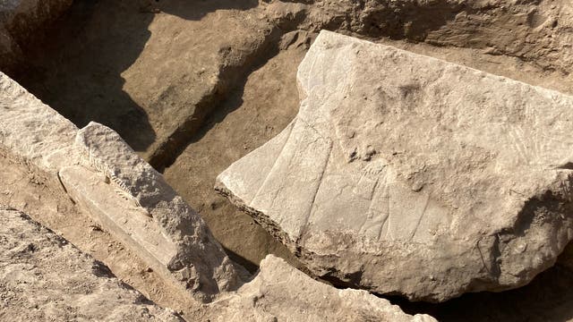 Eine archäologische Ausgrabungsstätte mit frei gelegten, großen Steinen und Erdschichten. Die Oberfläche der Steine zeigt feine Linien und Muster, die auf historische Gravuren hindeuten könnten. Die Szene ist von Sonnenlicht beleuchtet, das Schatten auf den Boden wirft.