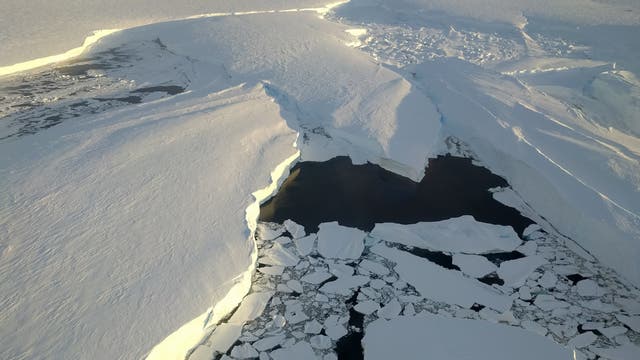Luftaufnahme einer riesigen, schneebedeckten Eisfläche in der Antarktis, die sich bis zum Horizont erstreckt. In der Mitte des Bildes ist ein großer Riss zu sehen, der mit dunklem Wasser gefüllt ist und von zerbrochenen Eisschollen umgeben ist. Die Szene wird von der tiefstehenden Sonne beleuchtet, die lange Schatten auf das Eis wirft.