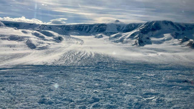 Eine weite, verschneite Landschaft mit einem Gletscher, der sich durch ein Tal zwischen schneebedeckten Bergen erstreckt. Der Himmel ist bewölkt, und Sonnenstrahlen brechen durch die Wolken, was der Szene eine dramatische Beleuchtung verleiht. Die Oberfläche des Gletschers zeigt Risse und Unebenheiten, die die Bewegung des Eises andeuten.
