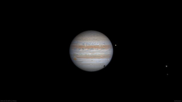 Das Bild zeigt den Planeten Jupiter mit seinen charakteristischen Wolkenbändern in verschiedenen Brauntönen und dem Großen Roten Fleck. Mehrere seiner Monde sind als kleine, helle Punkte sichtbar, die den Planeten umgeben. Der Hintergrund ist schwarz.