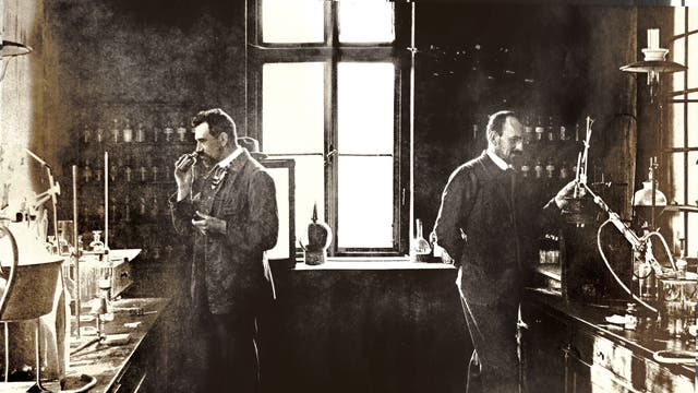 Zwei Männer in einem historischen Labor, umgeben von Glasflaschen und chemischen Apparaturen. Einer der Männer hält ein Objekt in der Hand und scheint es zu untersuchen, während der andere an einem Tisch arbeitet. Im Hintergrund sind Regale mit weiteren Laborutensilien zu sehen. Das Bild vermittelt eine Atmosphäre wissenschaftlicher Forschung aus einer früheren Zeit.