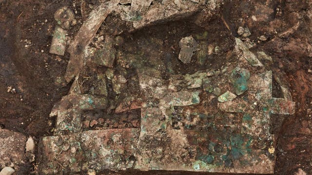 Eine archäologische Ausgrabungsstätte zeigt ein antikes, stark korrodiertes Metallobjekt im Erdreich. Um das Objekt herum sind goldene Schmuckstücke und blaue Perlen verstreut.