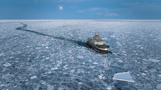 Ein Eisbrecher fährt durch eine weite, mit Eisschollen bedeckte Meeresfläche. Der Himmel ist bewölkt, und der Mond ist sichtbar. Das Schiff hinterlässt eine schmale, eisfreie Spur im Wasser.