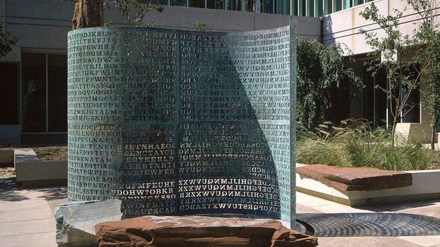 Eine Skulptur im Freien, bestehend aus einer großen, gebogenen Metallplatte mit eingravierten Buchstabenreihen, die wie ein Code oder Rätsel wirken. Die Skulptur steht auf einem Platz vor einem modernen Gebäude mit Glasfenstern. Um die Skulptur herum sind Bäume und Pflanzen zu sehen, die eine harmonische Verbindung zwischen Kunst und Natur schaffen. Die Buchstaben werfen Schatten auf den Boden, was dem Kunstwerk eine zusätzliche Dimension verleiht.