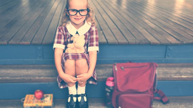 Ein junges Mädchen mit großer Brille sitzt mit angezogenen Beinen auf einer Treppe. Neben ihr steht eine rote Schultasche.