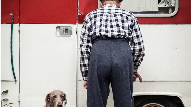 Ein Mann steht vor einem Wohnwagen. Neben ihm sitzt ein Hund und blickt in die Kamera.