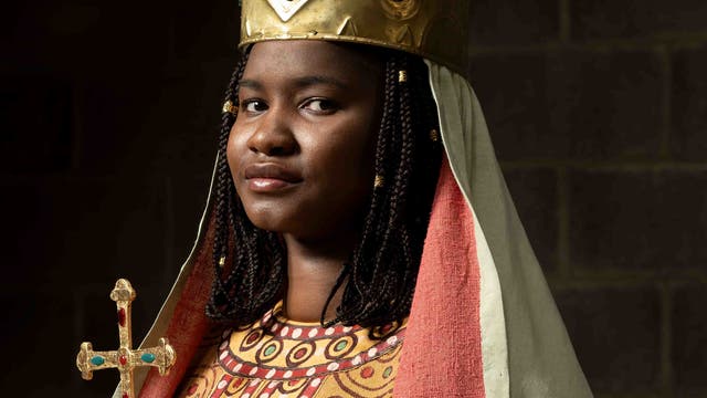 Eine Person in traditioneller Kleidung mit einer goldenen Krone und einem Umhang. Sie hält ein verziertes Kreuz. Der Hintergrund ist dunkel, was den Fokus auf die Person lenkt.