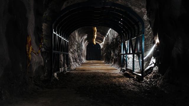Ein beleuchteter Tunnel mit stützenden Metallbögen und einer unebenen, erdigen Oberfläche. An den Wänden sind einige farbige Markierungen zu sehen. Der Tunnel verläuft geradeaus und ist von dunklen, felsigen Wänden umgeben. Oben verläuft ein Rohr entlang der Decke.