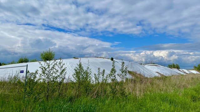 Eine weitläufige Landschaft mit grünen Wiesen und Büschen im Vordergrund. Im Hintergrund sind große, mit weißen Planen abgedeckte Hügel zu sehen, die möglicherweise landwirtschaftliche Materialien schützen. Der Himmel ist teils bewölkt mit blauen Abschnitten. Ein Zaun mit einem kleinen blauen Schild ist links im Bild sichtbar.