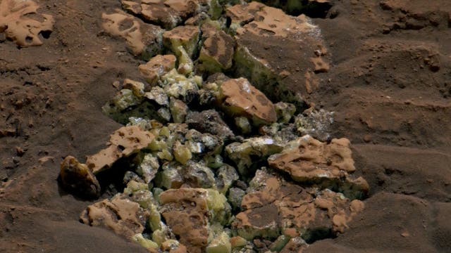 Ein zertrümmerter bräunlicher Gesteinsbrocken mit gelben Kristallen zwischen den Fragmenten.