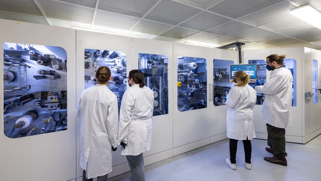 Eine Gruppe von Personen in weißen Laborkitteln steht vor einer großen, modernen Maschine in einem Labor. Die Maschine hat mehrere Fenster, durch die komplexe mechanische Teile sichtbar sind. Eine Person bedient ein Touchscreen-Display an der Maschine. Die Szene vermittelt eine professionelle, wissenschaftliche Umgebung.