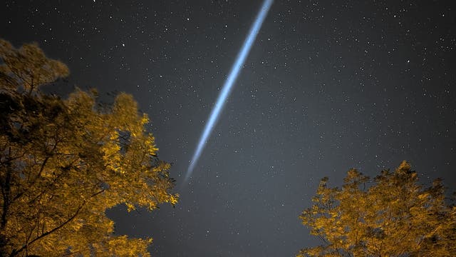 Ein klarer Nachthimmel mit zahlreichen Sternen. Ein heller, diagonaler Lichtstrahl durchquert das Bild von oben links nach unten rechts. Im Vordergrund sind die Silhouetten von Bäumen zu sehen, die von unten beleuchtet werden. Der Lichtstrahl könnte auf ein astronomisches Phänomen wie einen Meteor oder Kometen hinweisen.