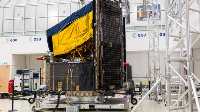 Ein Satellit in einem Labor der Europäischen Weltraumorganisation (ESA). Der Satellit ist mit goldener und schwarzer Folie bedeckt und steht auf einer Plattform, umgeben von technischen Geräten und Kabeln. Im Hintergrund sind ESA-Logos und große Fenster zu sehen.