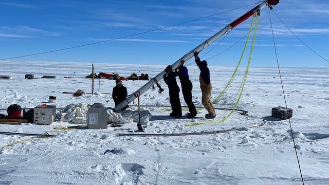 Eine Gruppe von Personen arbeitet in einer verschneiten, eisigen Landschaft der Antarktis daran, einen großen Eisbohrer aufzustellen. Der Himmel ist klar und blau. Um sie herum sind verschiedene Ausrüstungsgegenstände und Kisten auf dem Boden verteilt. Im Hintergrund sind weitere Strukturen oder Fahrzeuge zu sehen. Die Szene vermittelt den Eindruck einer wissenschaftlichen Expedition in einer abgelegenen, kalten Umgebung.