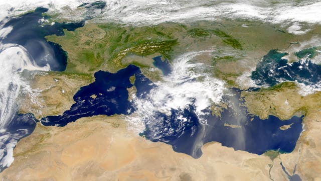 Satellitenbild des Mittelmeers, das auf der Aufnahme blau erscheint. Die umliegenden Kontinente Afrika, Asien und Europa erscheinen in Grün- und Brauntönen. Über Italien in der Bildmitte ist ein großer Wolkenkomplex zu sehen