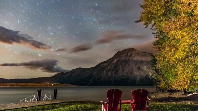 Rote Stühle laden in Kanada an historischen Stätten und in Nationalparks zum Verweilen und Ausruhen ein. Hier schweift der Blick über den Waterton-See und die ersten Herbststernbilder, die im Osten aufgehen. Das Scheinwerferlicht eines vorbeifahrenden Autos erhellte den Vordergrund. 
