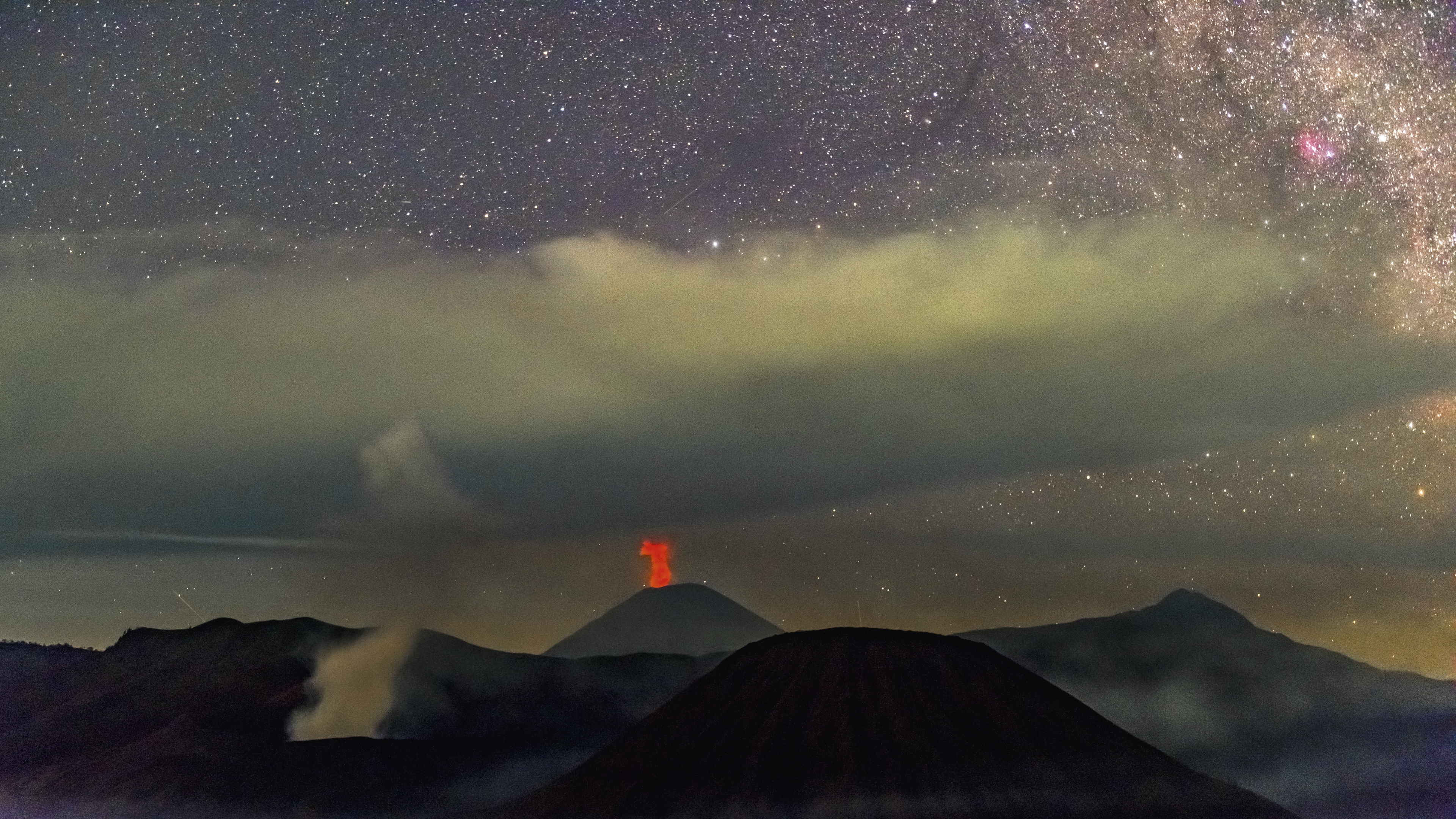 Der rauchende Krater des Vulkans Bromo und der Feuer speiende Semeru, der höchste Berg auf der Insel Java, bilden eine exotische Kulisse vor den Wolkenfronten der irdischen Atmosphäre und den interstellaren Wolken in der Milchstraße.