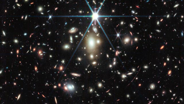 Eine Aufnahme des Weltraumteleskops, die eine Vielzahl von Galaxien in unterschiedlichen Formen und Farben zeigt, verteilt über ein dunkles, sternenübersätes Universum. Im Zentrum leuchtet ein heller Stern mit auffälligen Lichtstrahlen. Die Galaxien variieren in Größe und Helligkeit, einige erscheinen spiralförmig, andere elliptisch. Das Bild vermittelt die Weite und Vielfalt des Universums.