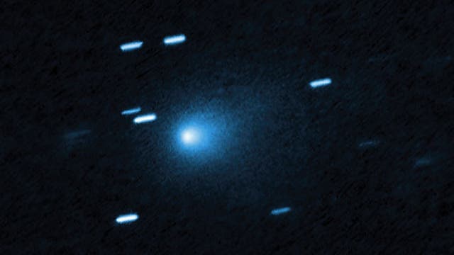 Ein astronomisches Bild zeigt einen Kometen im Weltraum, der von einem hellen, leuchtenden Kern und einem diffusen Schweif umgeben ist. Mehrere Sterne erscheinen als längliche Lichtspuren im Hintergrund, was auf die Bewegung des Kometen während der Aufnahme hinweist. Der dunkle Hintergrund hebt die bläuliche Färbung des Kometen hervor. Das Bild vermittelt den Eindruck von Bewegung und Tiefe im Universum.
