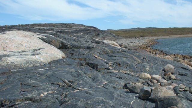 Eine felsige Küstenlandschaft mit dunklen, geschichteten Gesteinsformationen im Vordergrund. Der Himmel ist leicht bewölkt, und rechts im Bild ist ein ruhiges Gewässer zu sehen. Die Landschaft erstreckt sich in die Ferne, wobei das Ufer von kleineren Steinen gesäumt ist.