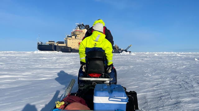 Blick vom Anhänger eines Schneemobils nach forne auf die Fahrer und im Hintergrund ein Schiff im Eis.
