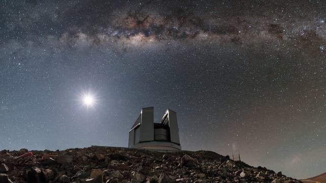 Ein Observatorium steht auf einem felsigen Hügel unter einem klaren Nachthimmel. Die Milchstraße erstreckt sich horizontal über das Bild, während ein heller Himmelskörper, vermutlich der Mond, links leuchtet. Der Himmel ist mit zahlreichen Sternen übersät, die eine beeindruckende Kulisse für das Observatorium bieten.