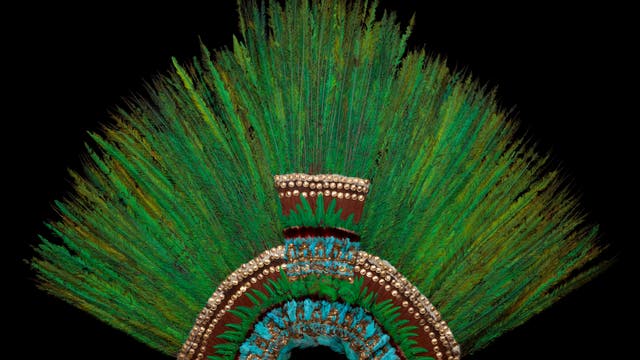 Ein traditioneller Kopfschmuck aus grünen und blauen Federn, verziert mit goldenen Elementen, auf schwarzem Hintergrund. Die Federn sind fächerförmig angeordnet und bilden ein kunstvolles Muster.