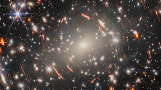 Ein beeindruckendes Bild des Weltraums, das eine Vielzahl von Galaxien zeigt, die in verschiedenen Farben leuchten, darunter helle Weiß- und Orangetöne. Im Zentrum des Bildes befindet sich eine große, helle Galaxie, umgeben von vielen kleineren, die einen tiefen, dunklen Hintergrund ausfüllen. Einige Sterne erscheinen als helle Lichtpunkte mit strahlenden Lichtspitzen. Dieses Bild vermittelt die Weite und Schönheit des Universums.