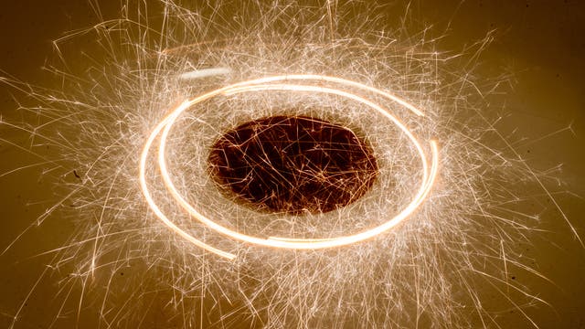 Brennende Wunderkerzen, die tangential an einer Scheibe aufgehängt sind, hinterlassen sprühende Funken, während sich die Scheibe im Kreis dreht. Eine lange Belichtungsdauer macht die glühenden Stäbe als Strichspuren sichtbar.