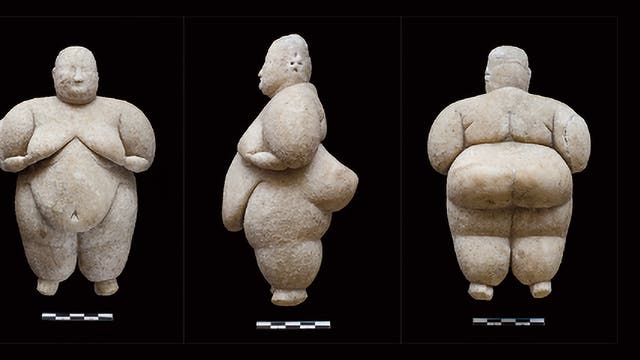Dreifache Ansicht einer prähistorischen »Venusfigurine« aus Stein, die eine füllige weibliche Form darstellt. Die Figur ist aus drei Perspektiven zu sehen: vorne, seitlich und hinten. Jede Ansicht zeigt die detaillierte Gestaltung der Figur, einschließlich der betonten Rundungen und der schlichten Gesichtszüge. Unter jeder Ansicht befindet sich eine Maßstabsleiste. Die Figur steht vor einem schwarzen Hintergrund.