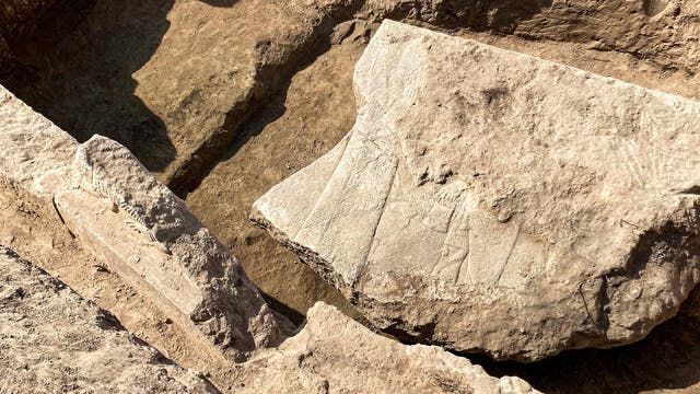 Eine archäologische Ausgrabungsstätte mit frei gelegten, großen Steinen und Erdschichten. Die Oberfläche der Steine zeigt feine Linien und Muster, die auf historische Gravuren hindeuten könnten. Die Szene ist von Sonnenlicht beleuchtet, das Schatten auf den Boden wirft.