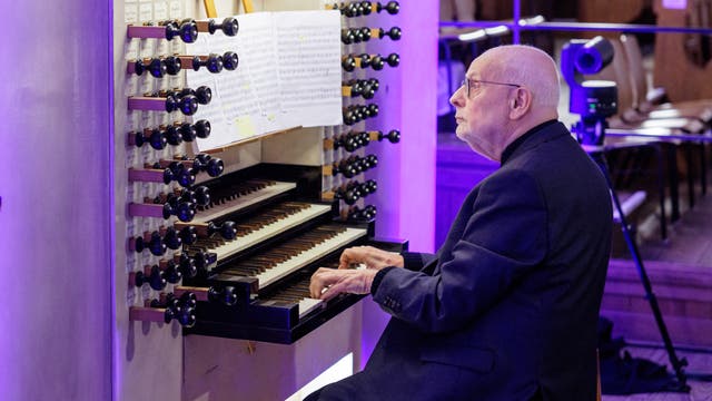 Ein älterer Mann spielt konzentriert auf einer großen Kirchenorgel. Die Orgel hat mehrere Manuale und zahlreiche Registerzüge. Notenblätter sind auf dem Notenständer platziert. Der Raum ist in violettes Licht getaucht, und im Hintergrund sind Kirchenbänke zu sehen.