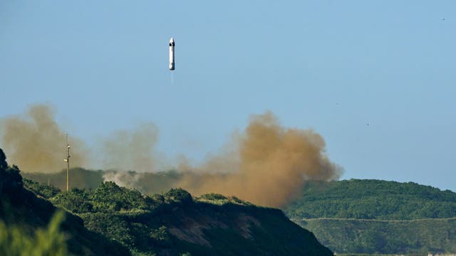 Eine Rakete steigt senkrecht in den blauen Himmel auf, während sie von einer grünen, bewaldeten Landschaft umgeben ist. Am Boden ist eine große Staubwolke zu sehen, die den Start begleitet. Im Hintergrund erstrecken sich Hügel.