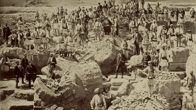 Eine historische Fotografie zeigt eine große Gruppe von Menschen, die auf und um große Felsblöcke und griechische Ruinen herumstehen. Viele Personen tragen traditionelle Kleidung und posieren für das Foto. Im Hintergrund ist eine hügelige Landschaft zu sehen. 