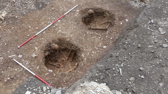 Zwei freigelegte menschliche Skelette in runden Gräbern auf einer archäologischen Ausgrabungsstätte. Die Skelette sind in gebeugter Position, umgeben von Erde. Zwei Messstäbe liegen parallel zu den Gräbern, um die Größe zu verdeutlichen. Im Hintergrund ist eine Steinmauer sichtbar.