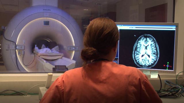 Ein Patient liegt im Hintergrund im MRT. Im Vordergrund schaut sich eine Frau im Kontrollraum die Ergebnisse auf einem Monitor an.