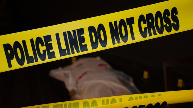 (amerikanisches) Polizei-Absperrband, dahinter schemenhaft eine 'Leiche' (vermutlich eine Schaufensterpuppe unter einem rot beschmierten weißen Laken).